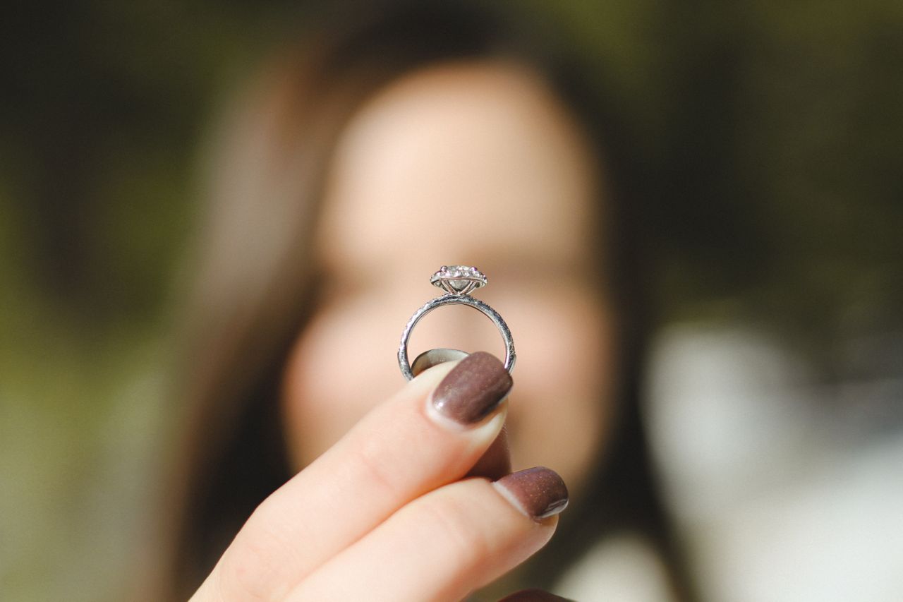 Engagement ring with intricate metalwork details seen from the side profile. The ring is held by a blurry figure in the background